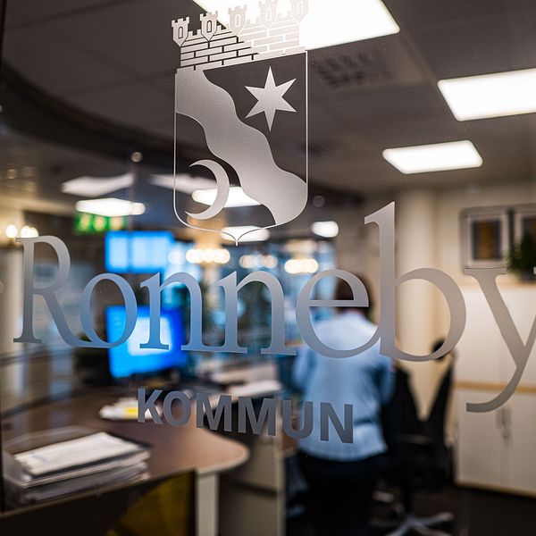ALT-text: Skylt med texten ”Ronneby kommun” på en glasdörr, med suddig bakgrund som visar ett kontor. En person i blå skjorta syns delvis i bakgrunden.