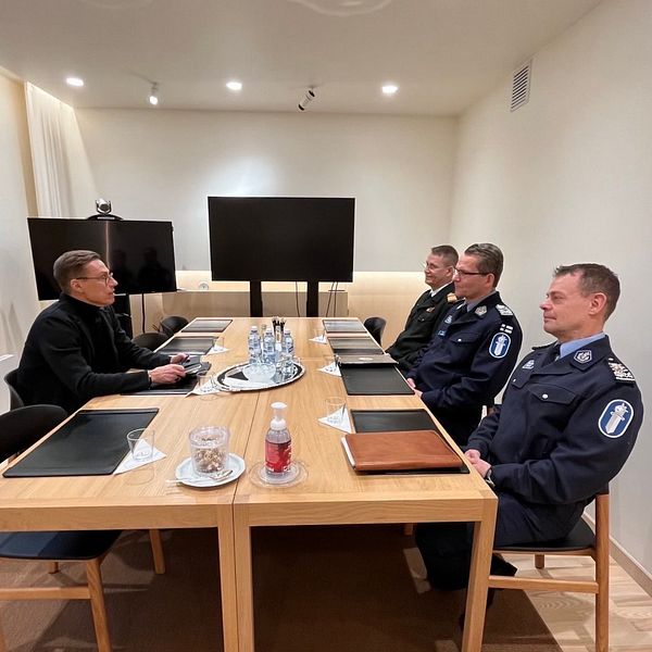 En konferensrumsscen med en träbord där en person i svart kläder sitter vid ena änden av bordet och arbetar på en laptop, medan tre uniformerade poliser sitter på andra sidan.