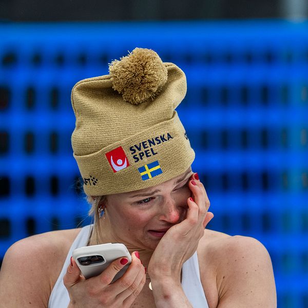 Person med ullmössa och telefon, blå bakgrund.