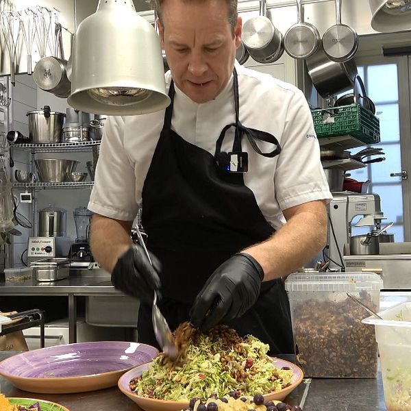 En kock i vit uniform och svarta handskar förbereder en stor sallad i ett restaurangkök. På bänken syns flera skålar med färgglada grönsaker och tillbehör, samt verktyg och utrustning för matlagning i bakgrunden.