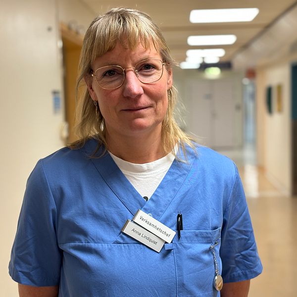 En sjuksköterska iklädd blå arbetskläder står i en lång korridor på ett sjukhus. Namnskylt och en penna syns i bröstfickan. Korridoren är ljus med vitt och blått som dominerande färger.