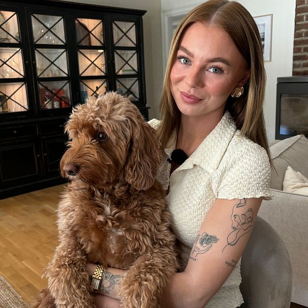 Kvinna med tatuerad arm sitter i ett vardagsrum och håller en brun hund i knät.