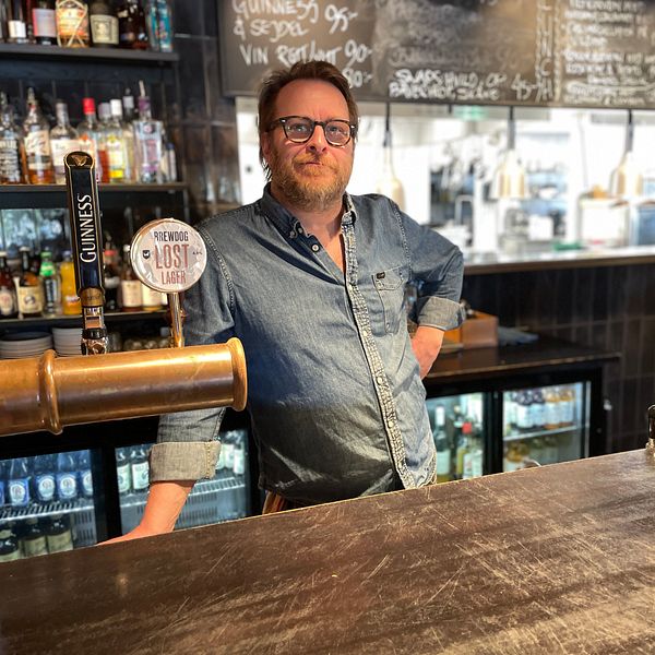 Bartender står bakom en bardisk med flaskor, ett ölkran och staplade tallrikar.