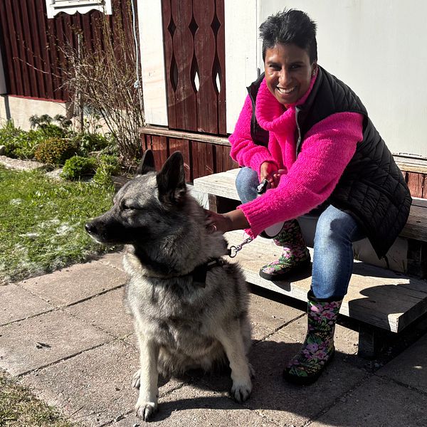 Person med rosa jacka sitter på trappa utanför hus med stor hund i koppel.