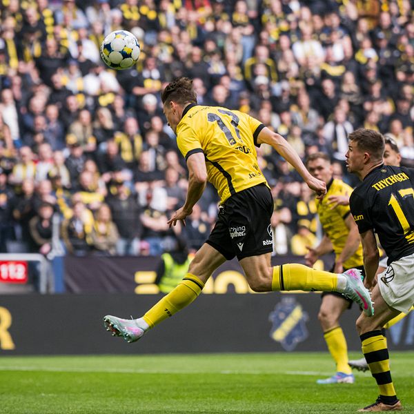 Fotbollsspelare i gult gör ett nickmål under match mot lag i svart framför stor publik.