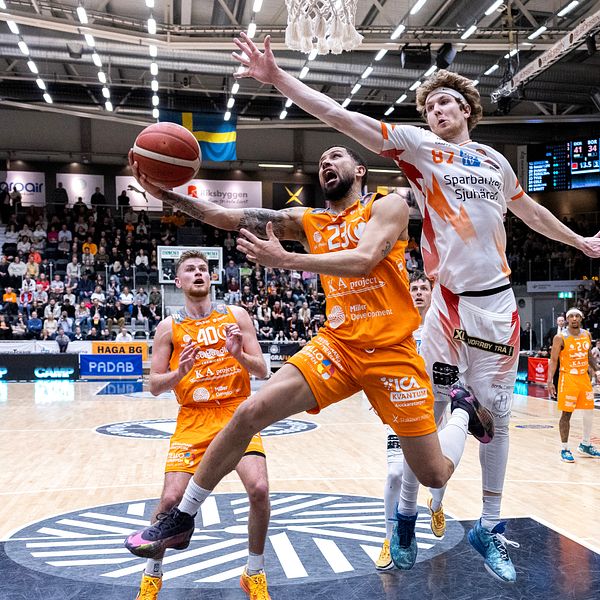 Basketspelare i orange och vitt hoppar mot korgen inför en fullsatt läktare.