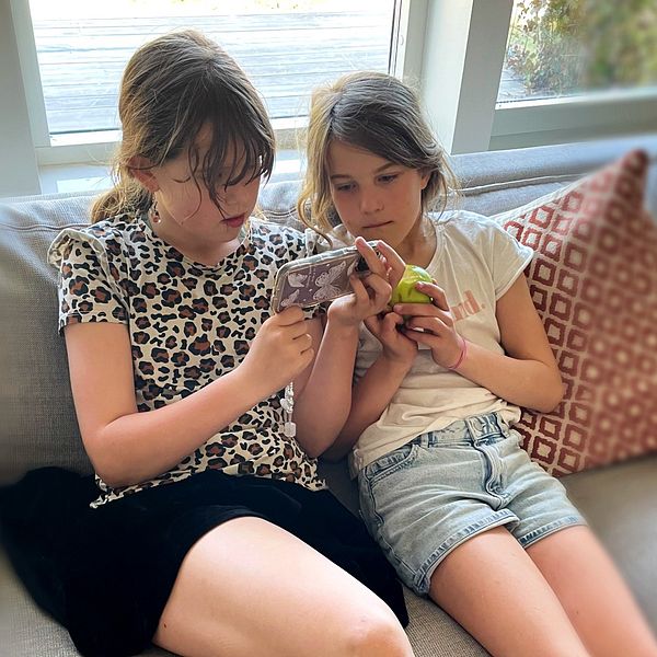 Två unga flickor sitter i soffan och tittar i en mobil tillsammans