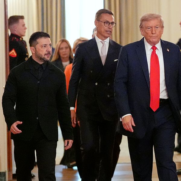 Volodymyr Zelenskyj går tillsammans med två män i kostym i en officiell byggnad.
