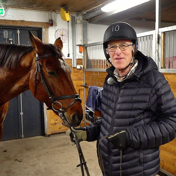 En person står i en häststall och håller i ett hästhuvud med en grimma. Personen bär en svart jacka och hjälm, medan stallinredningen i bakgrunden visar träväggar och stallfack. Det är en lugn miljö med inomhusbelysning.