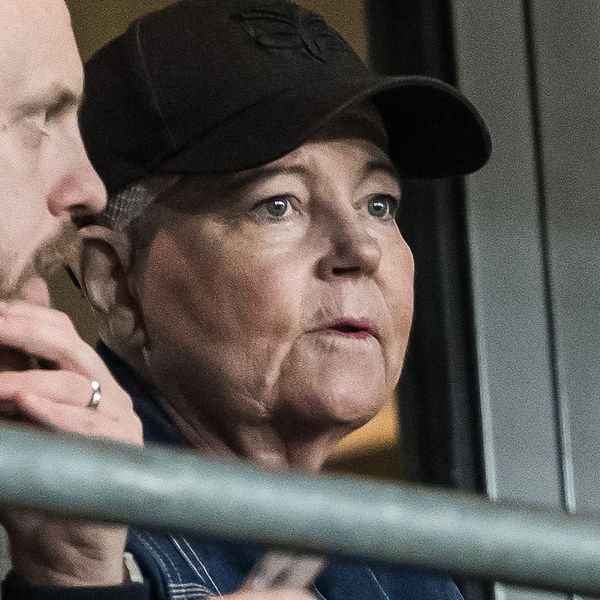 Två personer sitter i en läktare, den ena håller i en telefon och den andra ser mot en händelse i fjärran, med en stängsel i förgrunden.