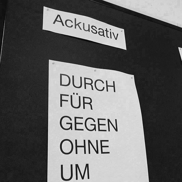 En svartvitt bild av en anslagstavla med tre rubriker: ”Ackusativ”, ”Dativ” och ”Befallning”. Under ”Ackusativ” listas prepositioner som ”DURCH”, ”FÜR”, ”GEGEN”, ”OHNE” och ”UM”. Under ”Dativ” finns prepositioner som ”AUS”, ”AUSSER”, ”BEI”, ”GEGENÜBER” och ”MIT”.