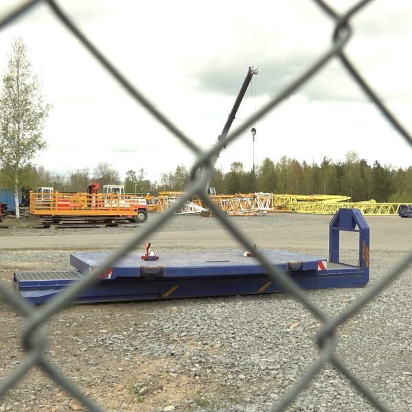 Industriområde sett genom stängsel, med maskiner och fordon på grusplan.