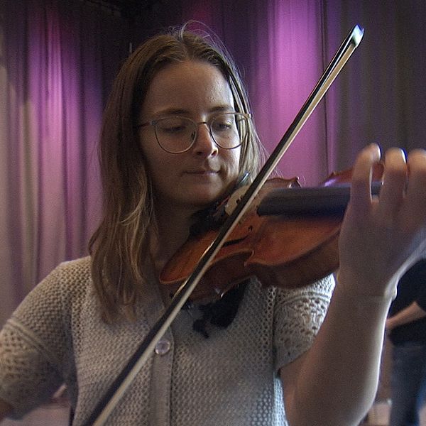 Ung person spelar fiol inomhus, omgiven av andra människor.