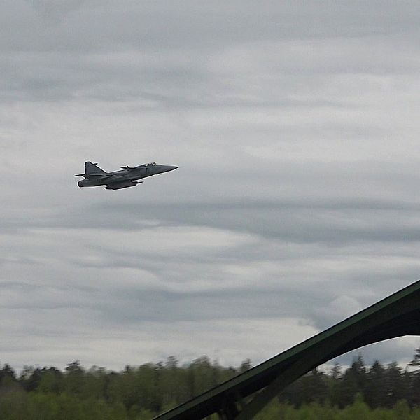 Stridsflygplan flyger lågt över skog och en militärhangar i förgrunden.