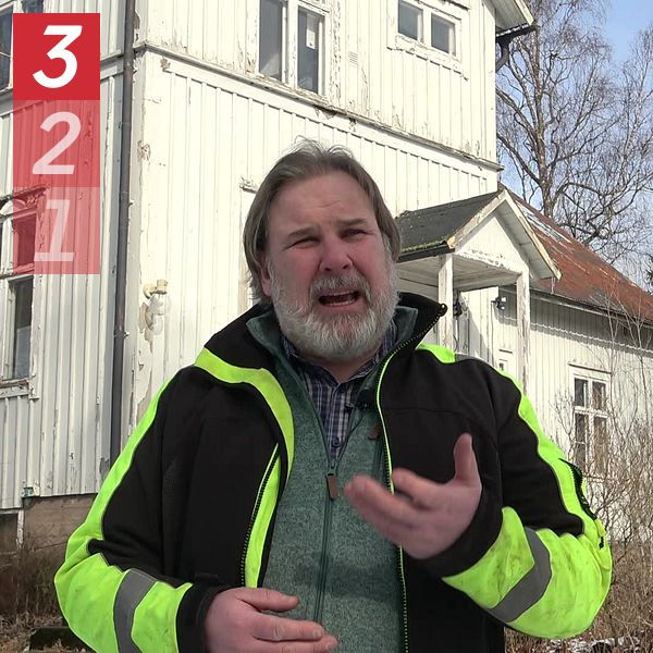 Person i gul varseljacka framför ett vitt trähus med natur i bakgrunden.