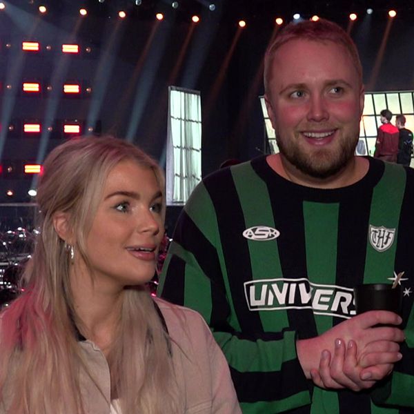 Programledarna Kristina ”Keyyo” Petrushina och Edvin Törnblom står framför en upplyst scen. I bakgrunden syns även stolsrader, rekvisita och strålkastare.