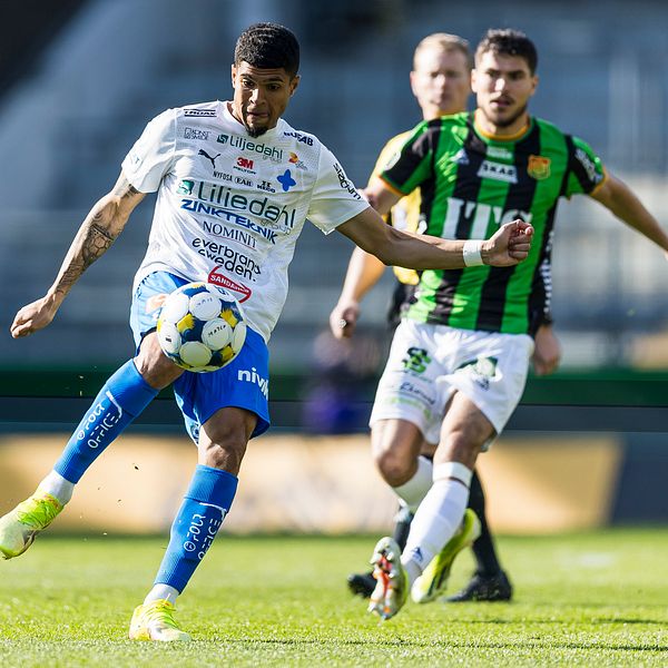 Fotbollsspelare i vit och blå dräkt sparkar bollen, motståndare i grönsvart förföljer.
