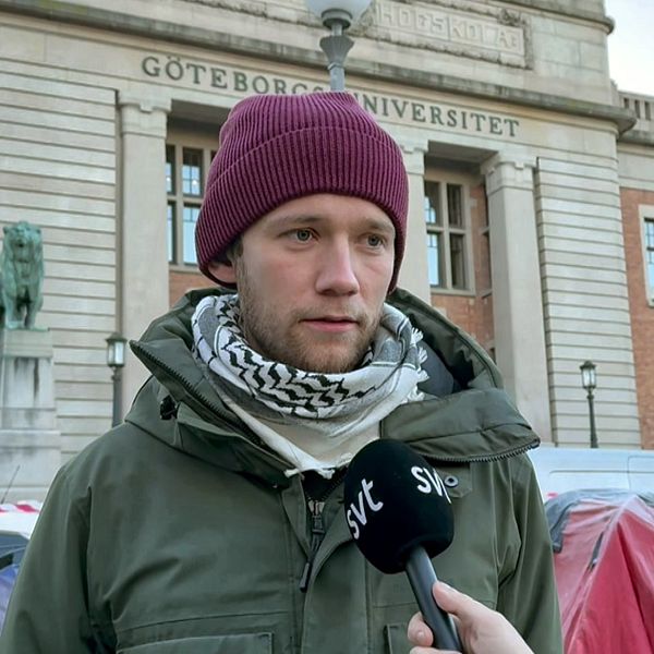 En person i en grön jacka och röd mössa intervjuas framför Göteborgs universitet. Byggnadens fasad är synlig i bakgrunden, inklusive universitetets namn. Intervjupersonen bär även en scarf över halsen och en journalist med mikrofon ställer frågor. I bakgrunden syns även tält och ett avspärrningsband.