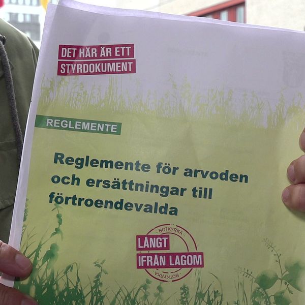 En person håller upp ett dokument med titeln ”Reglemente för arvoden och ersättningar till förtroendevalda”. Dokumentet är omslaget av grönt och gult med texten ”DET HÄR ÄR ETT STYRDOKUMENT” tydligt synlig. Bakgrunden visar en byggnad och omgivande miljö.