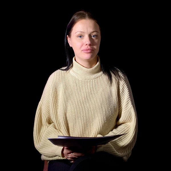 Stina sitter i en mörk miljö iförd en ljus stickad tröja och håller en som i sitt knä.