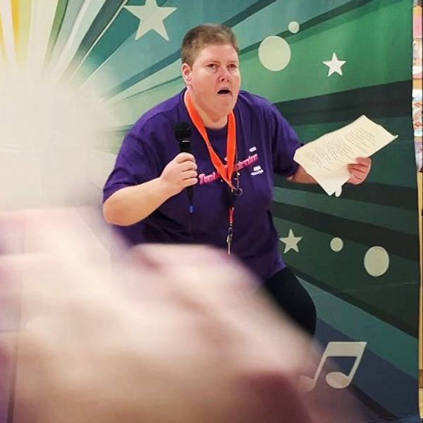 En person i lila tröja står framträdande på en scen med en färgglad bakgrund av stjärnor och musiknoter. Personen håller ett papper och gestikulerar med entusiasmerande rörelser. Framför scenen syns händer som klappar, vilket tyder på publikens uppmuntran. I bakgrunden finns en kontorsmiljö med stolar och bord.