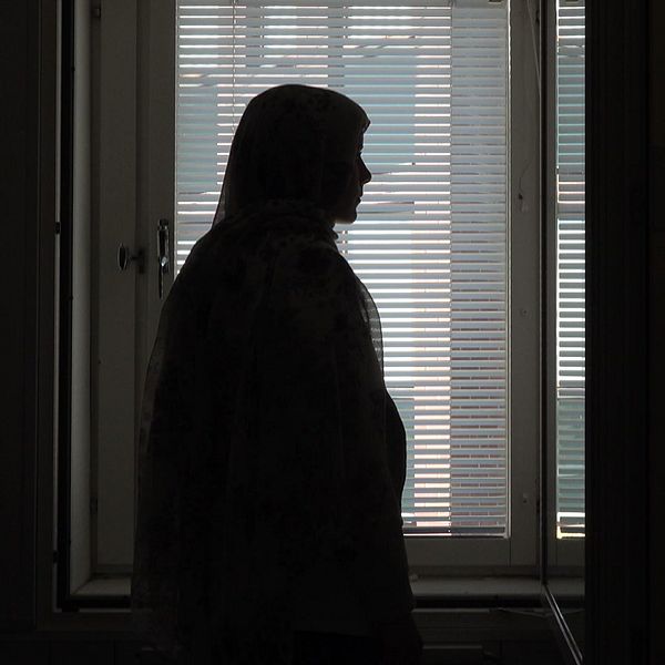 En kvinna i slöja står i profil mot ett fönster med neddragna persienner. Bilden är mörk och kvinnans ansikte syns inte