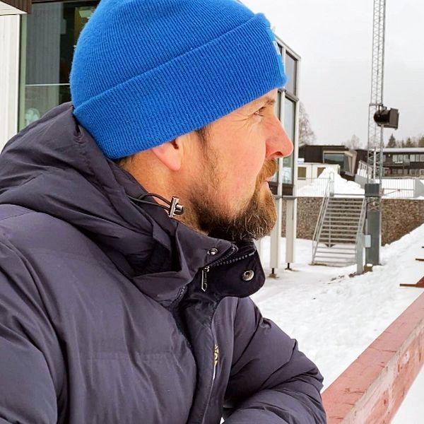 En person står utomhus iförd en blå mössa och mörk vinterjacka, med ryggen vänd mot kameran, och tittar ut över en snötäckt arena med kringliggande skog i bakgrunden.