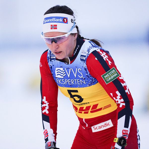En skidåkare i röd och gul tävlingsdräkt med nummer fem, står redo på snön, förberedd för en tävling. Dräkten har sponsormärken och är utrustad för kalla förhållanden.