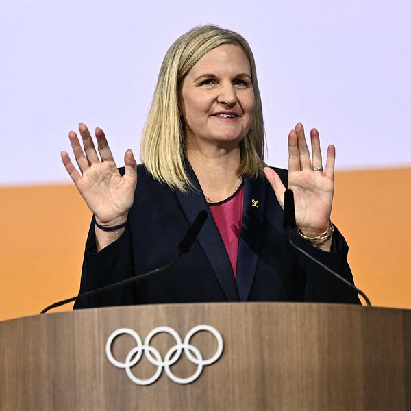 Kvinna talar vid olympisk podium med höjda händer.