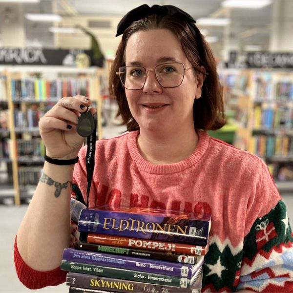 Personen håller en hög av böcker i vänster hand och en klocka i höger hand, klädd i en röd tröja med mönster. I bakgrunden syns bokhyllor i en biblioteksmiljö.
