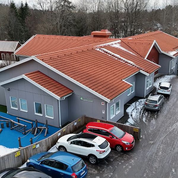 Vy över en skolbyggnad med röd takpannor, omgiven av en lekplats och en parkeringsplats med flera bilar. Lekplatsen har en blå yta och gungor, och i bakgrunden syns en skog med träd.
