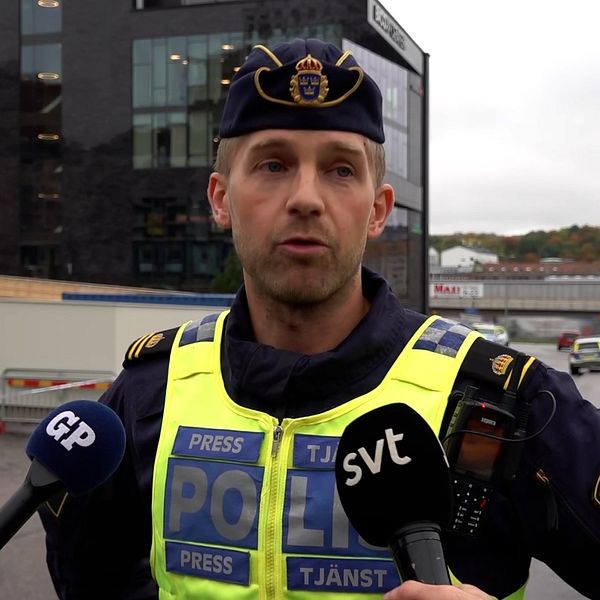 En polis står framför en byggnad och talar med journalister, som håller mikrofoner med logotyper från olika medier. Det är en mulen dag, och i bakgrunden syns en stadsmiljö med trafik och träd.