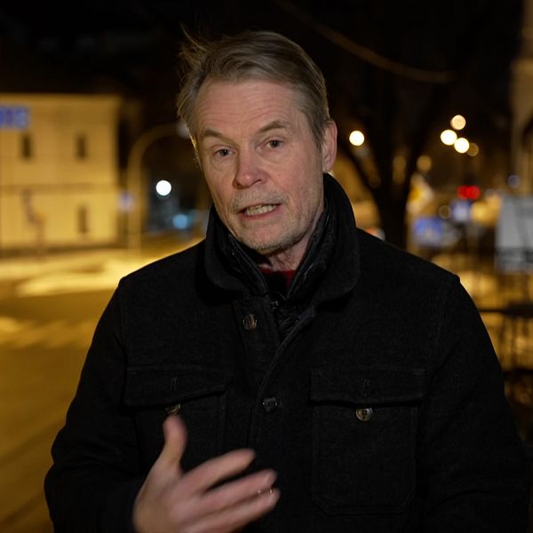 Person klädd i mörk jacka gestikulerar på en gata i en stad på kvällen.