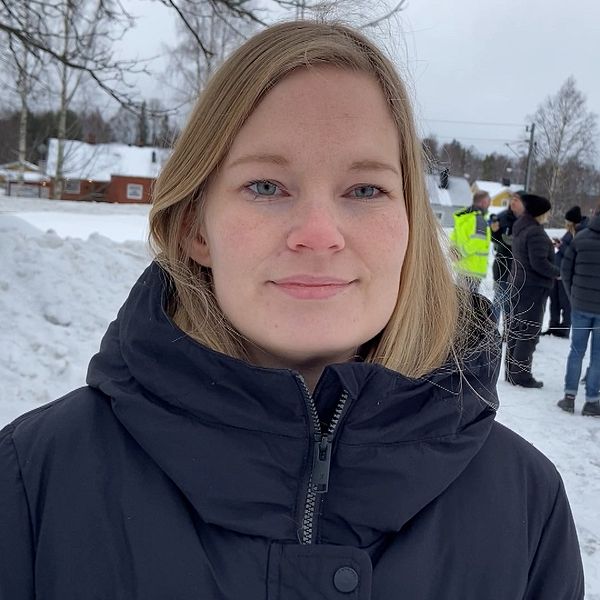 En vintermiljö med snö på marken. I bakgrunden samlas flera personer, några i neongula jackor, förmodligen del av ett arbetslag eller en grupp vid ett utomhusevenemang, framför en större snödriva omgiven av träd och byggnader.
