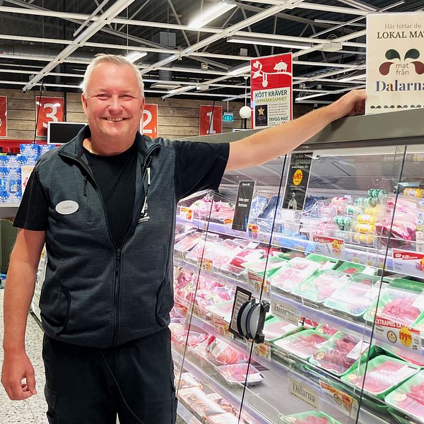 En icahandlare som står och ler vid köttdisken och pekar mot en skylt som det står lokalmat, Dalarna på.