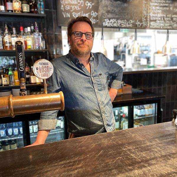 Bartender står bakom en bardisk med flaskor, ett ölkran och staplade tallrikar.