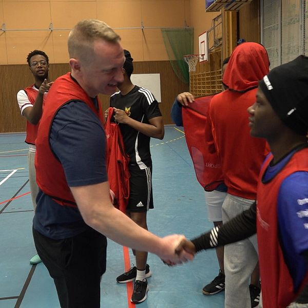 En grupp ungdomar i sportkläder befinner sig i en inomhusidrottshall. Politikerna Per Fredriksson skakar hand med en kille.