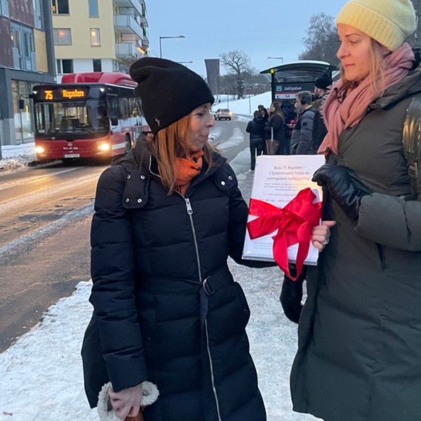 Två resenärer håller i en protestlista framför en busshållplats och en buss.