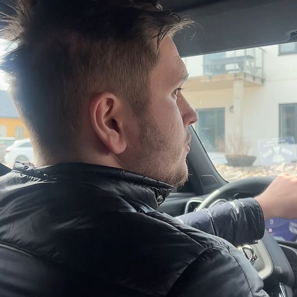 En person kör bil, hand på ratten. Byggnader syns utanför bilens fönster.