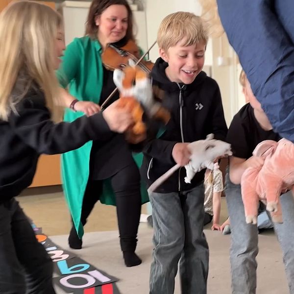 Barn leker med mjukisdjur medan en vuxen spelar fiol i bakgrunden.