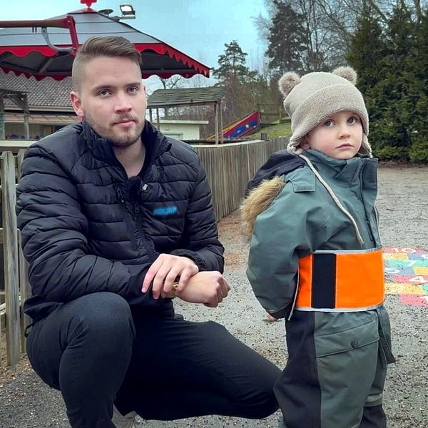 En vuxen och ett barn står vid en lekplats utomhus. Barnet bär en grön overall med en orange reflexväst och en mössa med öron. Lekplatsen har en färgglad lekställning i bakgrunden och omgivningen är grön och skuggigare med träd och gräsmatta.