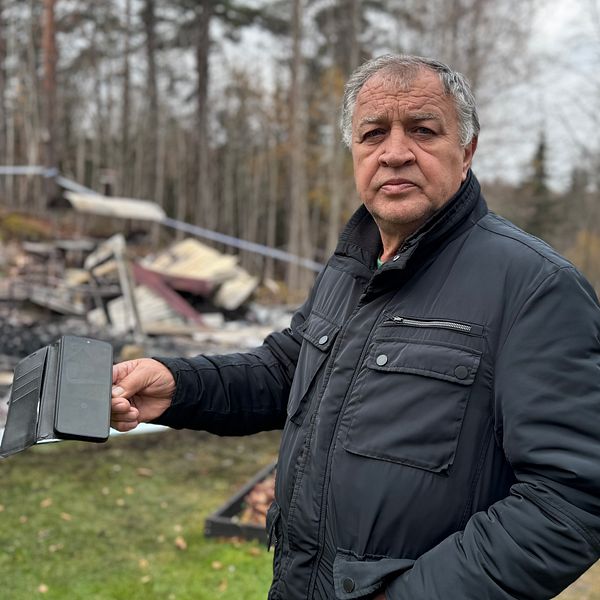En person i en svart jacka håller en bild på sin stuga och står framför en plats med nedbrunnet byggnadsrester. I bakgrunden syns en avspärrning och träd, vilket indikerar att området är efter en brand. Atmosfären är sorgsen och allvarlig.