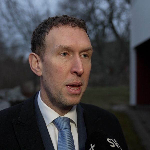 En man i kostym står utomhus i en dyster miljö med träd och en byggnad i bakgrunden. En mikrofon märkt ”SVT” syns framför.