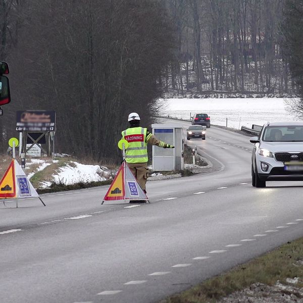 Väg 204, på vägen står en räddningstjänstbil och skyltar med texten olycka. En brandman gör tecken till trafiken att sänka hastigheten.