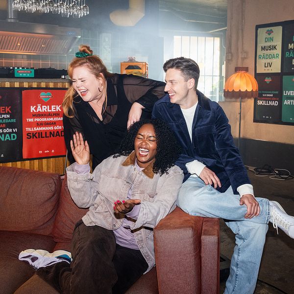 Linnea Wikblad, Emil Hansius och Assia Dahir sitter på en soffa i en informell miljö med affischer för Musikhjälpen i bakgrunden. Amie gestikulerar med handen, medan de andra två poserar avslappnat.