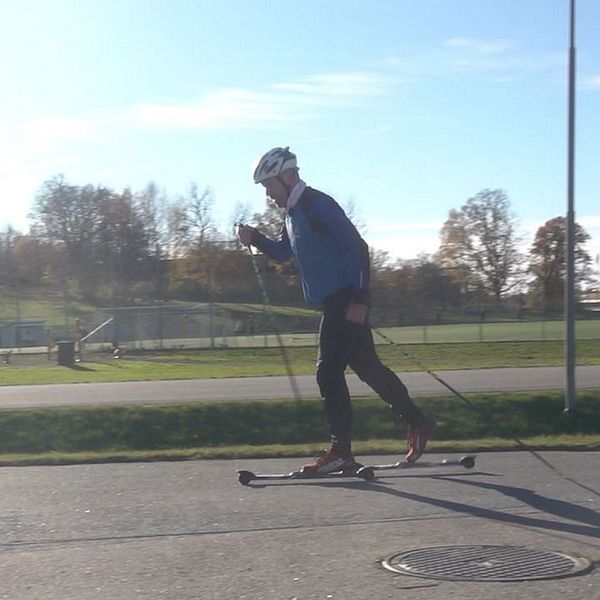 En person åker längdskidor på en asfaltväg under klar himmel. I vänster kant finns en nedräkning som visar siffrorna 3, 2, 1. I bakgrunden kan man se grönområden och sportanläggningar.