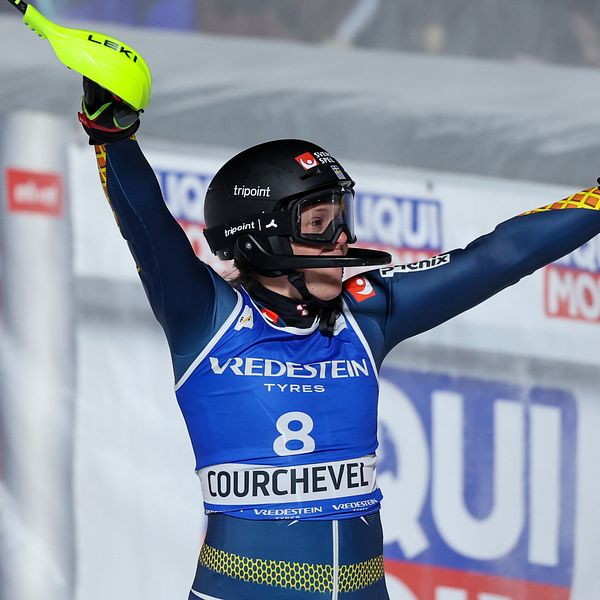 En alpin skidåkare i tävlingsdräkt firar med armarna i luften i Courchevel med startnummer 8. Reklambanderoller syns i bakgrunden.