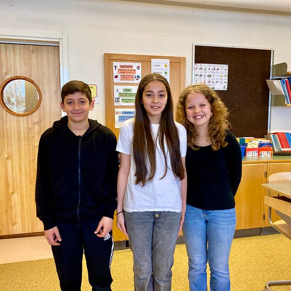 Tre elever står i ett klassrum, en kille till vänster, sedan två tjejer.