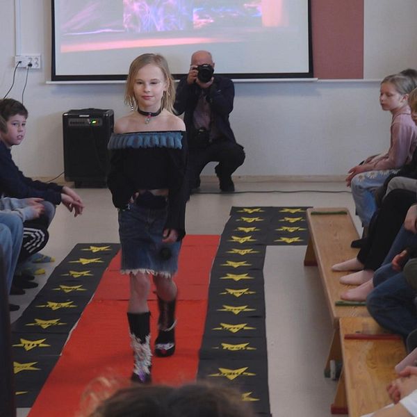 Ung modell går på röd catwalk i klassrum, åskådare sitter längs sidorna.