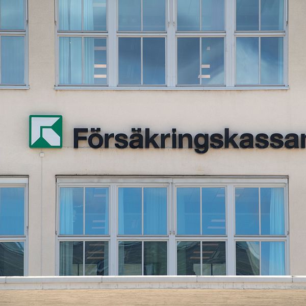 Skylt på en byggnad med texten ”Försäkringskassan” och en grön logotyp. Flera fönster syns i bakgrunden, vilket ger en modern kontorsmiljö.